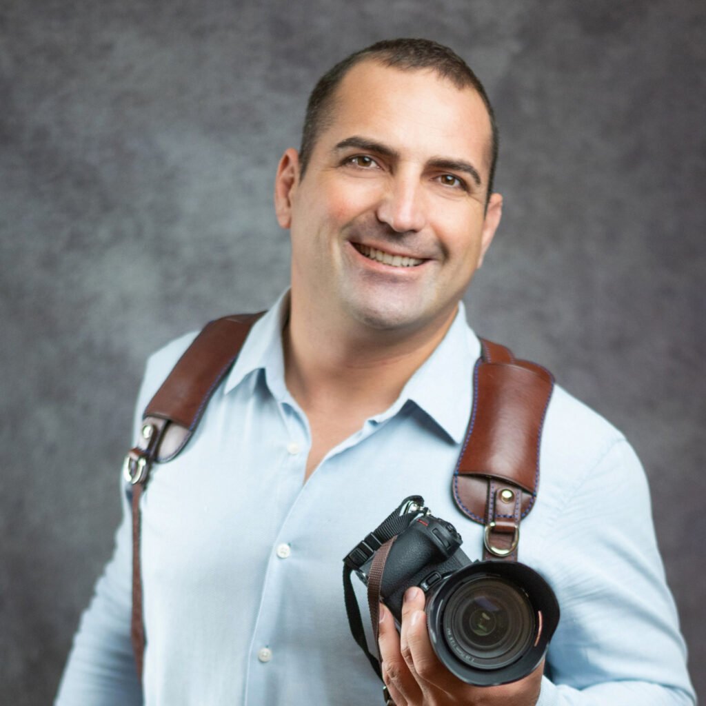 Dobó Imre, profi esküvői fotós mosolyogva tartja kezében professzionális fényképezőgépét. Világoskék inget és barna bőr vállhevedert visel, amely elegáns és praktikus kiegészítője egy esküvői fotográfus felszerelésének. A háttér egy letisztult, szürke stúdiófelület, amely kiemeli Imre barátságos és magabiztos kisugárzását. Esküvői fényképészként a legfontosabb pillanatokat örökíti meg, természetes és érzelemdús képeken keresztül.