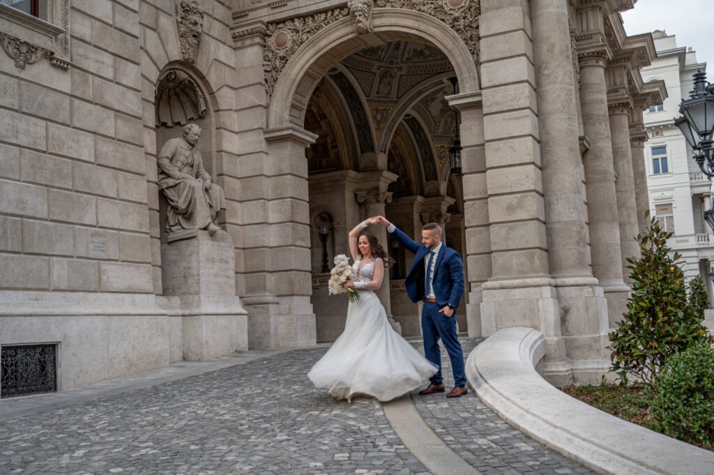 Esküvői fényképész által készített romantikus pillanat Budapesten – a menyasszony és a vőlegény egy történelmi épület árkádjai alatt táncol. A menyasszony kecsesen pörög, miközben a vőlegény gyengéden vezeti őt a kockaköves úton.