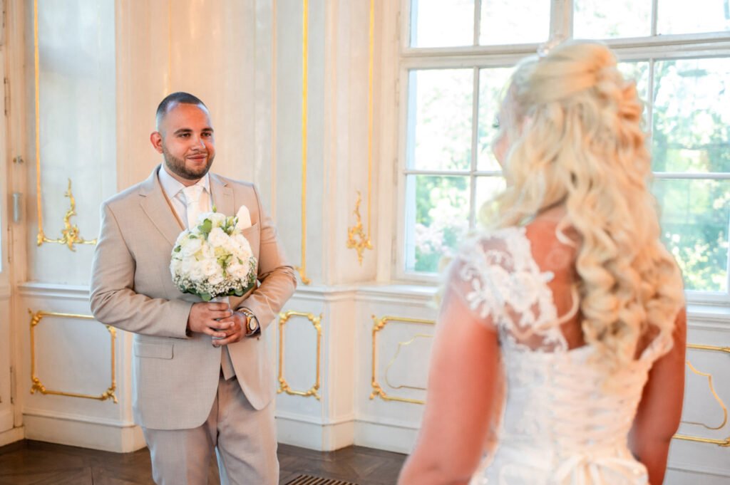 A vőlegény boldog mosollyal nézi menyasszonyát, miközben kezében egy fehér rózsákból és zöldekből álló esküvői csokrot tart. A menyasszony csipkés hátú, klasszikus menyasszonyi ruhájában áll előtte, hosszú szőke fürtjei lágyan omlanak vállára. A Gödöllői Királyi Kastély fényűző aranyozott falai és hatalmas ablakai romantikus, elegáns hátteret biztosítanak ennek a megható pillanatnak. Egy profi esküvői fotográfus által megörökített érzelemdús jelenet, amely a szerelmesek első találkozásának varázsát idézi fel a nagy napon.