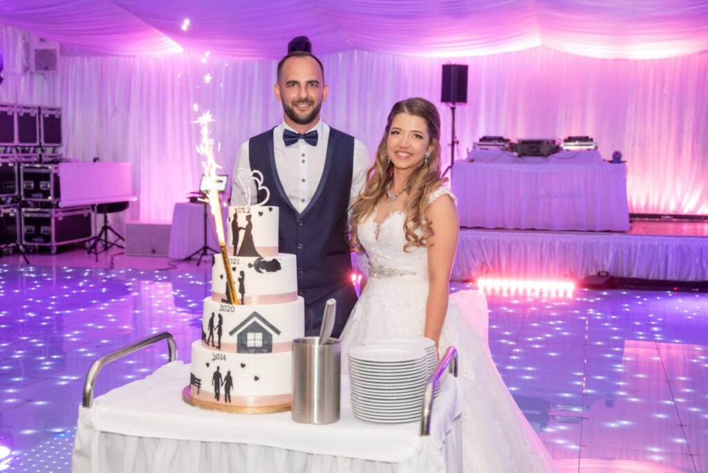 A menyasszony és a vőlegény boldogan állnak az esküvői torta mellett, amely egyedi dizájnnal és csillagszóróval díszített. A torta oldalán a pár közös életének fontos dátumai és szimbólumai láthatók. A menyasszony elegáns csipkés esküvői ruhát visel, míg a vőlegény klasszikus sötétkék öltönyben és csokornyakkendőben áll mellette. A világító táncparkett és a háttérben lévő DJ pult ünnepi hangulatot teremt. Esküvői tortavágás fotózás, ünnepi menyasszonyi pillanat, kreatív esküvőfotó, prémium esküvői fényképezés.