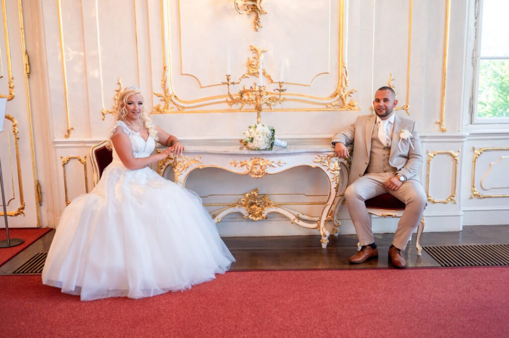 Egy elegáns menyasszony és vőlegény egy kastély pompás báltermében, aranyozott faldekorációval és klasszikus bútorokkal körülvéve. A menyasszony hercegnői stílusú esküvői ruhában egy antik széken ül, míg a vőlegény bézs öltönyben, lazán, magabiztosan helyezkedik el vele szemben. A köztük lévő márványasztalon a menyasszony fehér virágcsokra pihen, két elegáns gyertya mellett. Az esküvői fényképezés itt különleges és időtálló hangulatot kap, tökéletes választás azok számára, akik kastélyi esküvői fotókat keresnek egy profi esküvői fotográfus szemén keresztül.