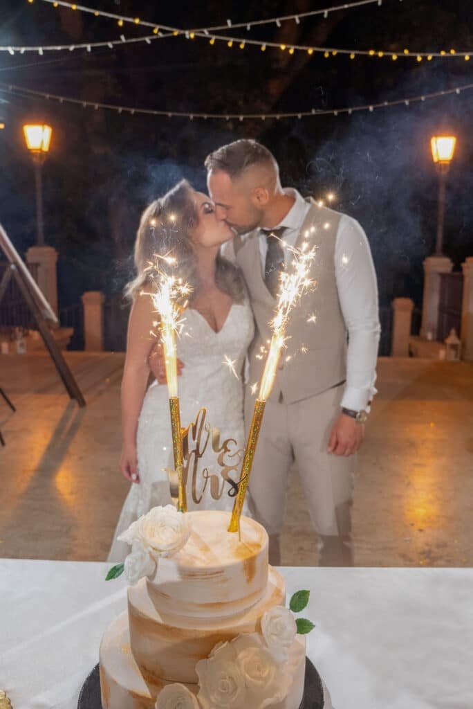 Egy romantikus esküvői pillanat, ahol a menyasszony és a vőlegény szenvedélyesen csókolózik a díszes esküvői torta előtt. A tortán két arany csillagszóró ég, fénye beragyogja az esti kültéri helyszínt, ahol hangulatos fényfüzérek és utcai lámpák világítják meg az elegáns dekorációt. A menyasszony csipkés, fehér esküvői ruhában ragyog, míg a vőlegény világos bézs öltönyben áll mellette. Az esküvői fotográfus kreatívan örökítette meg a pillanatot, amely a fények, a romantika és az ünneplés tökéletes egyensúlyát mutatja.
