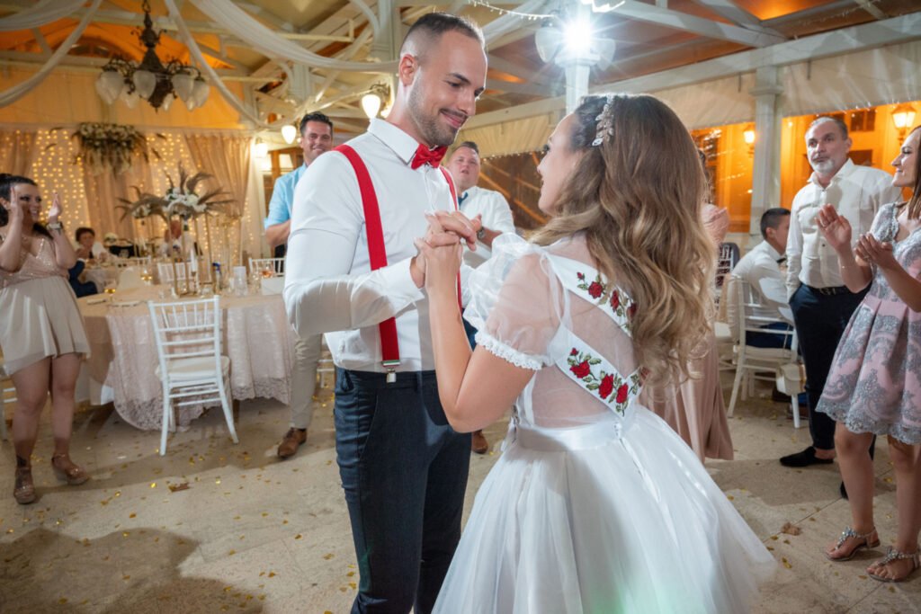 Esküvői fotográfia az esti mulatságon: a pár boldogan táncol a feldíszített sátorban, a menyasszony átöltözött egy piros hímzett, fehér ruhába, a vőlegény piros nadrágtartót és csokornyakkendőt visel, körülöttük vendégek tapsolnak.