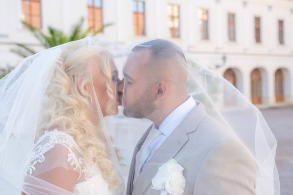 Egy romantikus esküvői fotó a Gödöllői Kastély elegáns udvarán, ahol a menyasszony és a vőlegény szerelmes csókot váltanak a fátyol alatt. A menyasszony csipkés esküvői ruhában, hosszú hullámos hajjal, a vőlegény pedig elegáns bézs öltönyben áll a történelmi kastély impozáns hátterében. Az esküvői fényképész tökéletesen megörökítette a meghitt pillanatot, amely a szerelmespár boldogságát és az esküvői fotózás varázsát sugározza.