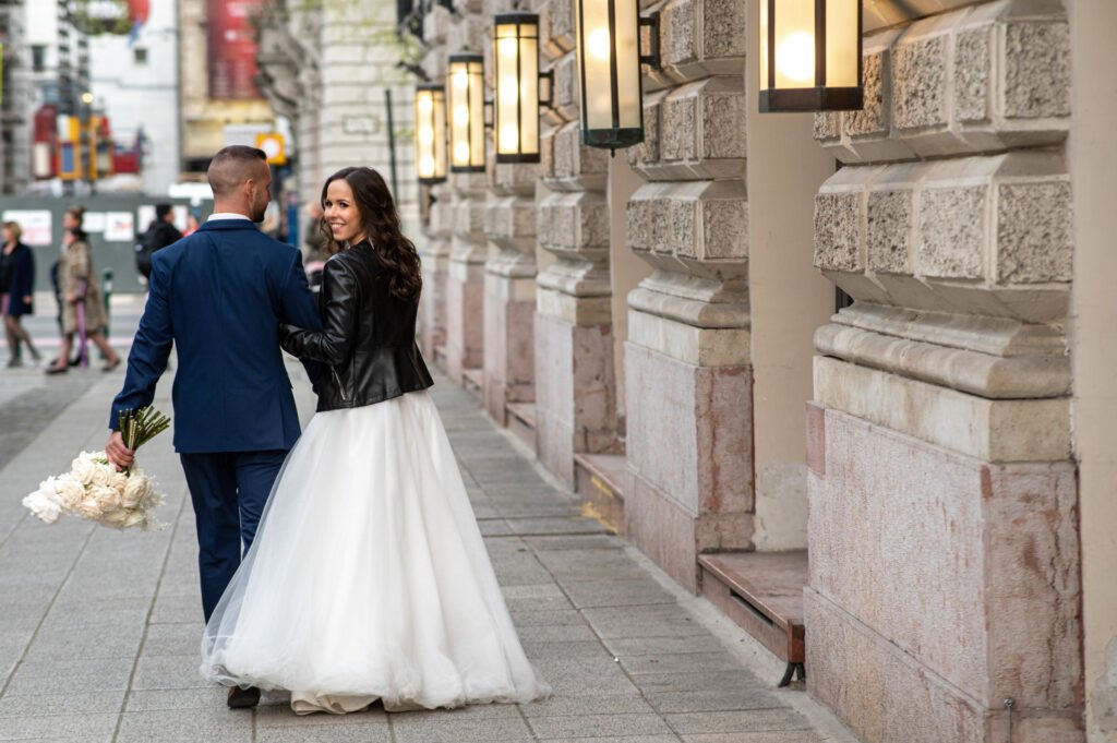 Esküvői fotós által készített kreatív városi kép egy friss házaspárról, akik elegáns, de lezser stílusban sétálnak a belvárosi épületek között. A menyasszony bőrdzsekit visel fehér esküvői ruhájához, a vőlegény pedig sötétkék öltönyben tartja kezében a virágcsokrot. Hangulatos utcai lámpák és történelmi épület díszíti a hátteret.