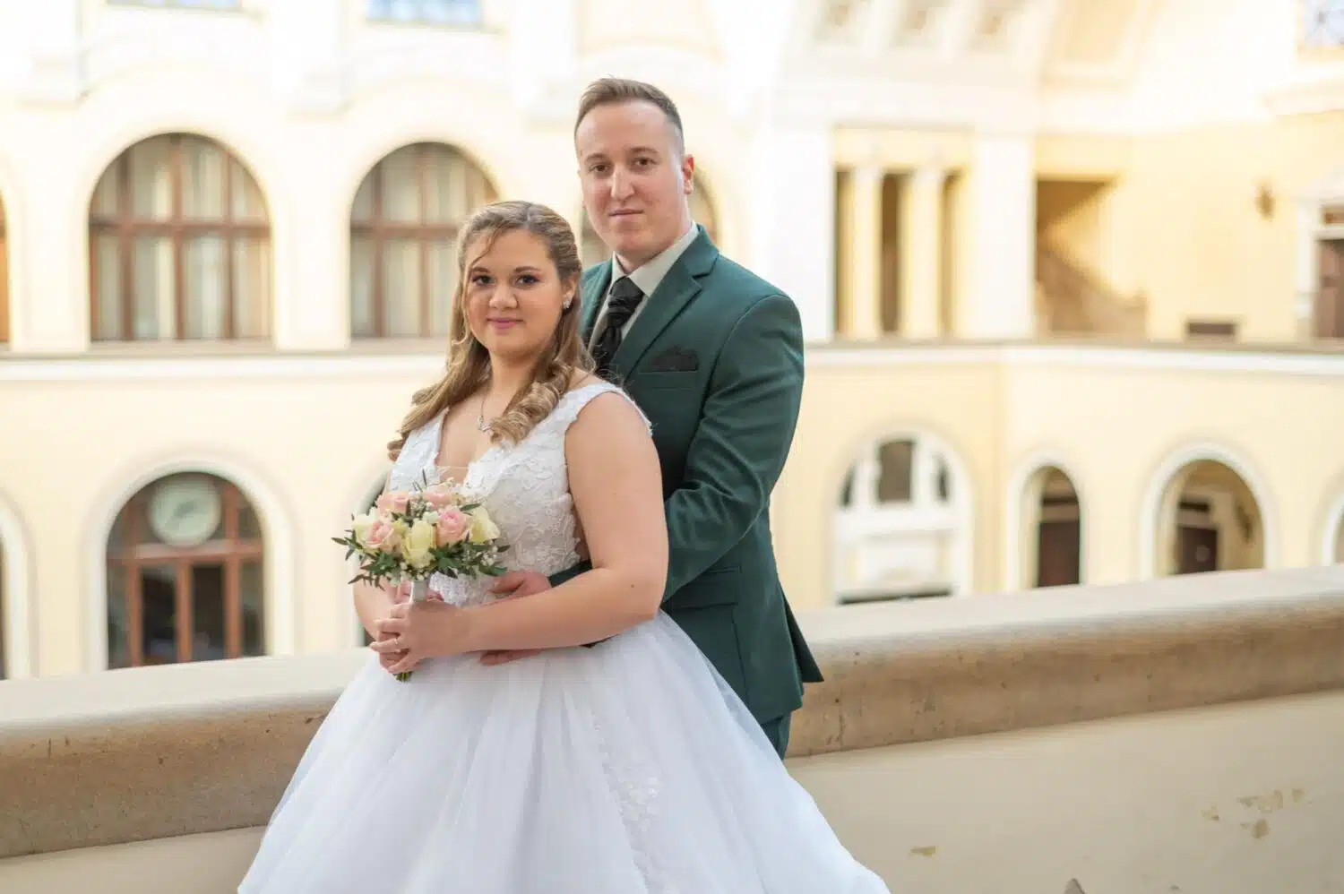 Egy boldog menyasszony és vőlegény elegáns esküvői fotója a Debreceni Egyetem lenyűgöző épületének teraszán. A menyasszony klasszikus fehér esküvői ruhában, kezében pasztellszínű rózsacsokorral áll, miközben a vőlegény stílusos zöld öltönyben gyengéden átöleli őt. A történelmi épület nagyívű ablakai és elegáns homlokzata impozáns hátteret biztosítanak, amely tökéletes helyszín egy profi esküvői fényképész számára. A kép a letisztult elegancia és az időtlen romantika tökéletes kombinációját nyújtja.