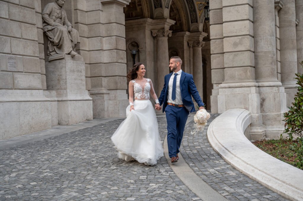 Esküvői fényképész által készített romantikus fotó Budapesten – a boldog pár kézen fogva sétál egy impozáns, oszlopokkal díszített épület előtt. A menyasszony elegáns, csipkés felsőrésszel és uszályos szoknyával tündököl, míg a vőlegény klasszikus sötétkék öltönyt visel.