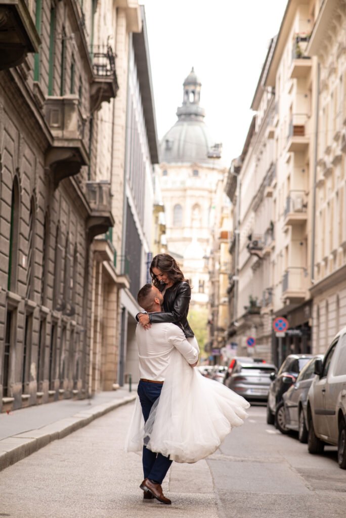 Esküvői fotográfus által készített romantikus kép egy menyasszonyról és vőlegényről, akik Budapest utcáin, a Szent István Bazilika előtt ölelik egymást. A menyasszony modern, fekete bőrdzsekit visel a fehér esküvői ruhájához, míg a vőlegény elegáns fehér ingben és sötétkék nadrágban emeli fel kedvesét a meghitt pillanatban.