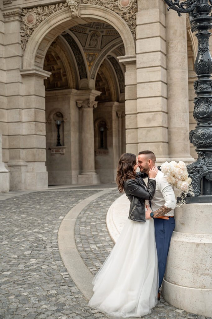 Esküvői fényképezés Budapest utcáin, ahol a menyasszony bőrkabátban és elegáns fehér ruhában áll vőlegényével egy klasszikus épület előtt. A fotográfus kreatív beállítással örökítette meg a pár romantikus pillanatát egy impozáns, boltíves háttérrel.