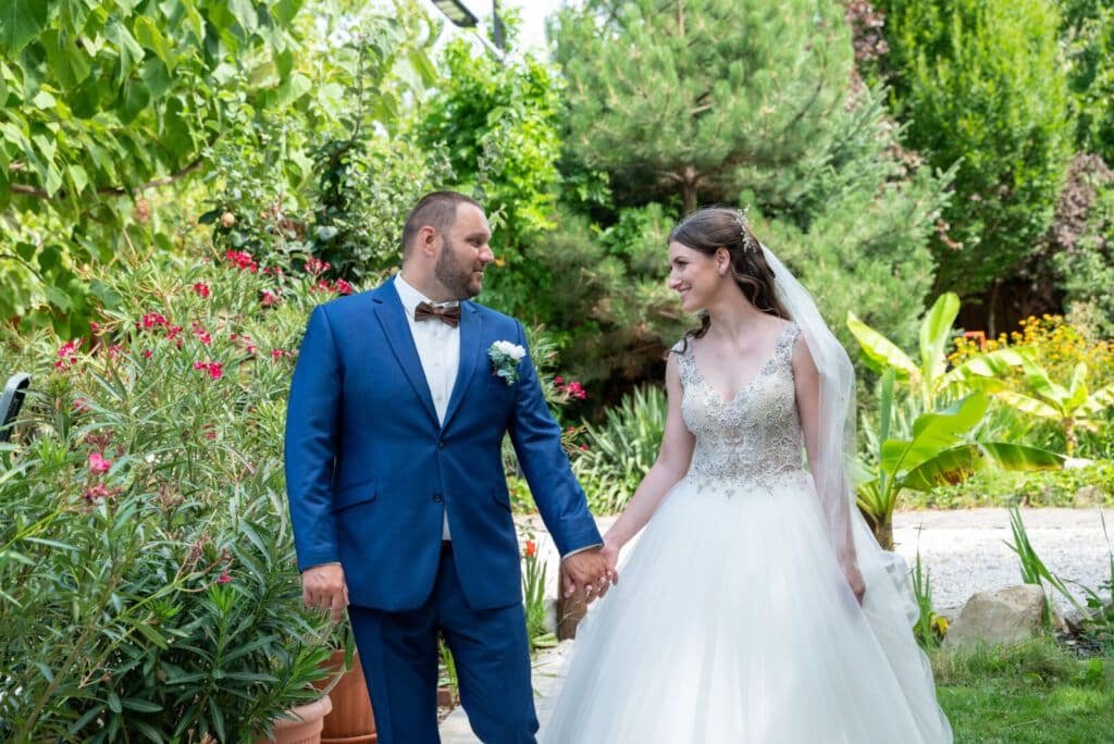 Egy boldog menyasszony és vőlegény kéz a kézben sétál egy gyönyörű, buja növényzettel teli kertben. A vőlegény elegáns kék öltönyt és barna csokornyakkendőt visel, míg a menyasszony egy lenyűgöző, csillogó felsőrésszel díszített fehér menyasszonyi ruhában ragyog. A háttérben színes virágok és egzotikus növények teszik még varázslatosabbá a helyszínt, amely tökéletesen illik egy természetközeli esküvői fotózáshoz. Ez az esküvői fotográfus számára inspiráló jelenet hűen megörökíti az örömteli és meghitt pillanatokat.