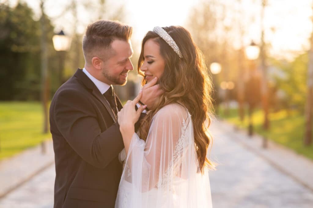 A nászünnep megörökítője által készített érzelemteli kép a Le Til Kúria fasorában naplementekor. A vőlegény gyengéden simogatja menyasszonya arcát, aki kristály fejdísszel és fátyollal díszített frizurával mosolyog, miközben a háttérben az alkonyi nap fénye szűrődik át a fák között.