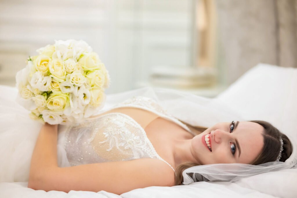 Esküvői fotóművész által készített intim boudoir portré: mosolygó menyasszony fehér csipkés fűzős ruhában, kristályos fejdísszel és tüll fátyollal, krémfehér és pezsgősárga rózsákból kötött kerek menyasszonyi csokorral, fehér párnákon pihenve, lágy természetes fényben.