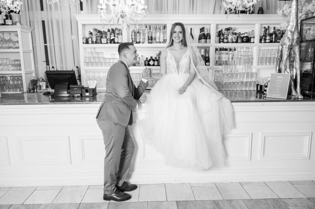 Játékos esküvői fotó a Le Til Kúria elegáns bárpultjánál, ahol a vőlegény szürke öltönyben térdel menyasszonya előtt, aki hercegnős szabású fehér ruhában a pulton ül. A háttérben kristálycsillárok és üvegpolcos bárszekrény látható.