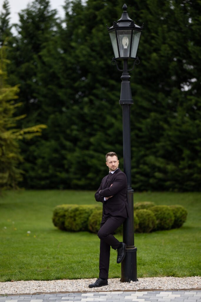 A nászriporter által készített stílusos kép a vőlegényről, aki klasszikus fekete öltönyben támaszkodik egy antik kandelábernek a Le Til Kúria gondozott parkjában, háttérben formára nyírt bokrokkal.