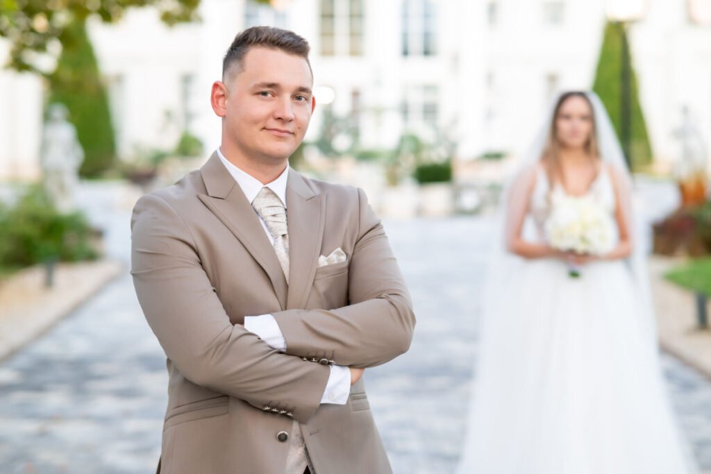 Egy magabiztos vőlegény áll a kamera előtt elegáns bézs színű öltönyben, keresztbe tett karokkal, miközben mosolyog. A háttérben kissé elmosódva a menyasszony látható hófehér esküvői ruhában, fátyollal és virágcsokorral a kezében. A helyszín egy elegáns kastélykert, macskaköves úttal, szobrokkal és gondozott növényekkel, amely exkluzív hátteret biztosít a romantikus esküvői fotózáshoz. Professzionális esküvőfotózás, amely kiemeli a vőlegény karakterét és a menyasszonnyal való meghitt kapcsolatát.