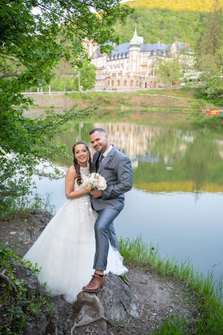 Esküvő fényképezés - Egy varázslatos esküvői fotózás a lillafüredi tó partján, ahol a boldog ifjú pár egy sziklás ösvényen áll a víz tükrében visszatükröződő kastély látványával a háttérben. A menyasszony gyönyörű, csipkével díszített hófehér esküvői ruhát visel, kezében pasztellszínű virágcsokorral. A vőlegény elegáns szürke öltönyben és barna bőrcipőben mosolyogva öleli őt. A környező fák és a tó nyugodt víztükre tökéletes harmóniát teremtenek a romantikus esküvői portré számára. Kreatív esküvőfotózás lenyűgöző természeti és építészeti környezetben.