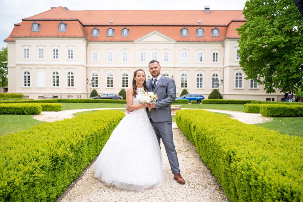 Egy gyönyörű esküvői fotó egy klasszikus kastély előkertjében, ahol a boldog ifjú pár mosolyogva áll a gondosan nyírt bokrok között. A menyasszony elegáns, csipkével díszített hófehér esküvői ruhát visel, kezében pasztellszínű virágcsokorral. A vőlegény szürke öltönyben, fehér ingben és barna bőrcipőben öleli őt. A háttérben a kastély impozáns homlokzata és a gondosan karbantartott park látható, amely időtálló eleganciát sugároz. Professzionális esküvői fotózás kastélyszálló romantikus környezetében, amely tökéletes választás a mesébe illő esküvői képekhez.