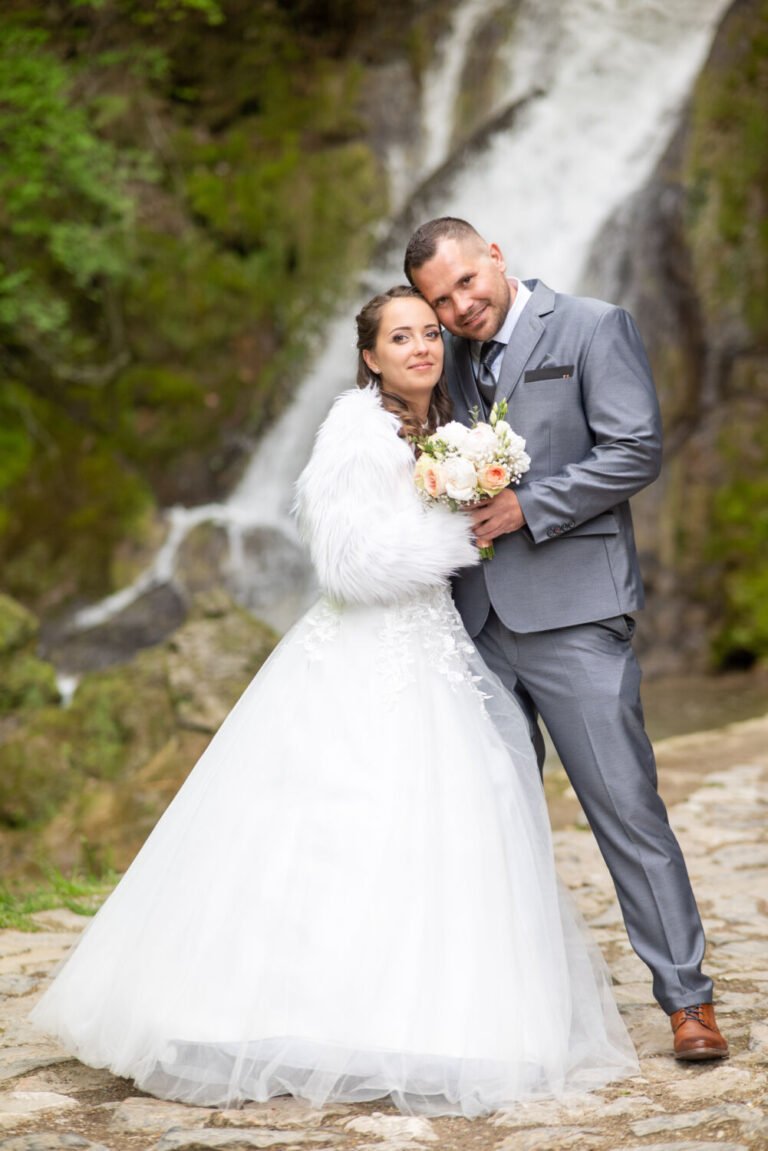 Egy boldog ifjú pár romantikus esküvői fotója a lillafüredi vízesés előtt. A menyasszony hófehér, tüllszoknyás esküvői ruhát és bolyhos fehér kabátot visel, kezében pasztellszínű virágcsokorral. A vőlegény elegáns szürke öltönyben, fehér ingben és barna bőrcipőben áll mellette, szeretetteljesen átölelve őt. A háttérben a vízesés zubogó vízsugarai és a mohával borított sziklák természetes, mesebeli hangulatot teremtenek. Professzionális esküvői fotográfia egy varázslatos helyszínen, amely időtálló emléket nyújt a szerelmeseknek.