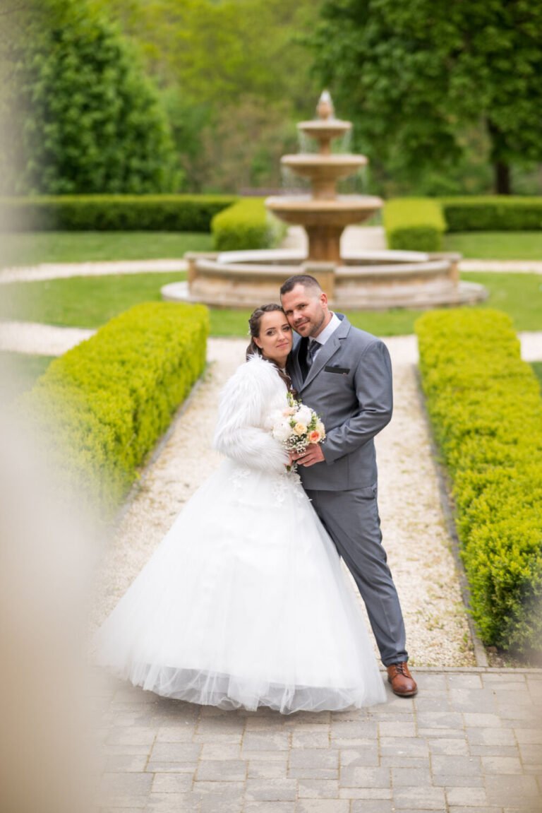 Esküvő fotózás - Egy romantikus esküvői portré egy gyönyörű kastélyparkban, ahol az ifjú pár egymáshoz bújva mosolyog a kamera felé. A menyasszony hófehér esküvői ruhát és bolyhos fehér kabátot visel, kezében pasztellszínű virágcsokorral. A vőlegény elegáns szürke öltönyben áll mellette, szeretetteljesen átölelve őt. A háttérben egy díszes kőszökőkút és gondosan nyírt bokrok adják a helyszín mesés atmoszféráját. A lágy előtér-homály kiemeli a pár meghitt pillanatát, amely tökéletesen tükrözi a nagy nap érzelmeit. Professzionális esküvőfotózás elegáns, romantikus környezetben.