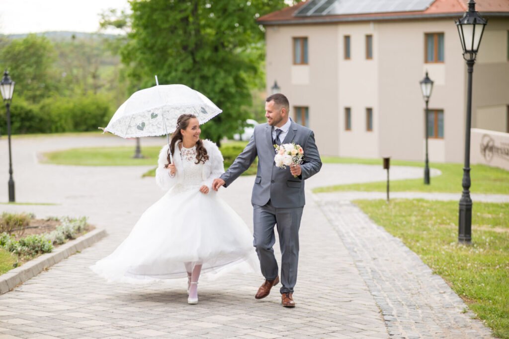 Egy boldog ifjú pár kézen fogva sétál egy macskaköves úton, mosolyogva nézve egymásra. A menyasszony csipkés, hófehér esküvői ruhát visel, bolyhos fehér kabáttal és egy díszes csipkeernyővel, amely romantikus hangulatot kölcsönöz a képnek. A vőlegény elegáns szürke öltönyben és barna bőrcipőben tartja a menyasszony virágcsokrát, amely pasztellszínű rózsákból és fehér virágokból áll. A háttérben modern épületek és vintage stílusú utcai lámpák láthatók, kiegészítve a helyszín elegáns atmoszféráját. Professzionális esküvőfotózás, amely természetes és meghitt pillanatokat örökít meg.