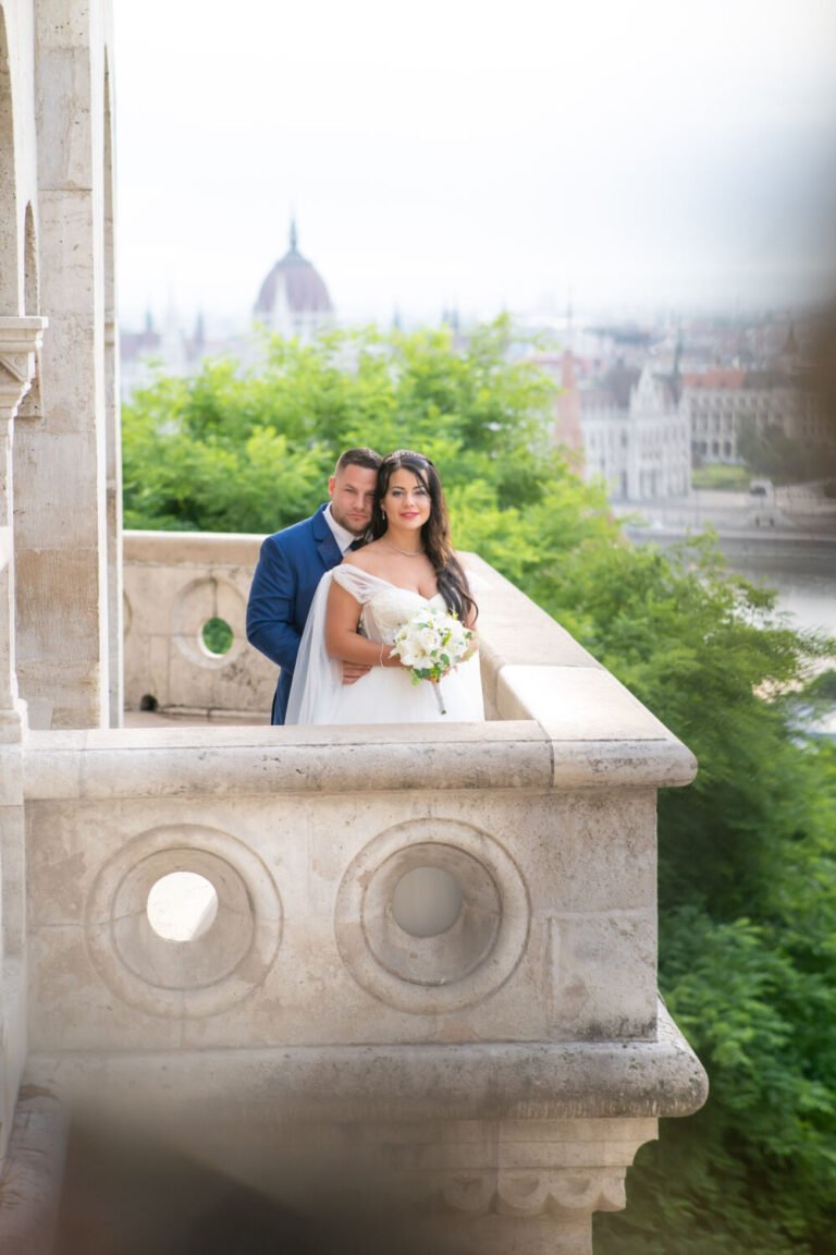 Esküvői kreatív fotós által megörökített légies pillanat: vállról lecsúszós fehér menyasszonyi ruhás menyasszony és királykék öltönyös vőlegény a Halászbástya jellegzetes kör motívumos kőkorlátjánál állnak. A menyasszony kezében fehér rózsacsokor, a háttérben a Parlament kupolája és zöldellő fák látszanak, miközben a pár szeretettel simul egymáshoz ebben a panorámás esküvői portréban.