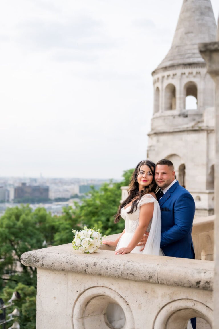 Esküvői portréfotós által megörökített meghitt pillanat: vállról lecsúszós fehér menyasszonyi ruhás, hosszú barna hajú menyasszony és királykék öltönyt viselő vőlegény a Halászbástya kőkorlátjánál állnak. A menyasszony kezében fehér rózsacsokor, a háttérben Budapest panorámája és a bástya jellegzetes fehér tornya látható, míg az előtérben a kör motívumos kőkorlát teszi még különlegesebbé ezt a romantikus kompozíciót.