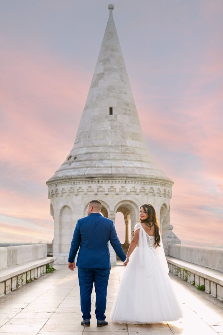 Esküvői fotós által megörökített varázslatos pillanat: kézen fogva sétáló ifjú pár a Halászbástya fehér tornya felé, ahol a menyasszony visszatekint válla felett. A fehér tüllruhás, hosszú barna hajú menyasszony és királykék öltönyös vőlegény sziluettje mögött rózsaszín-lilás alkonyi égbolt fest mesebeli hátteret ehhez a különleges esküvői portréhoz.