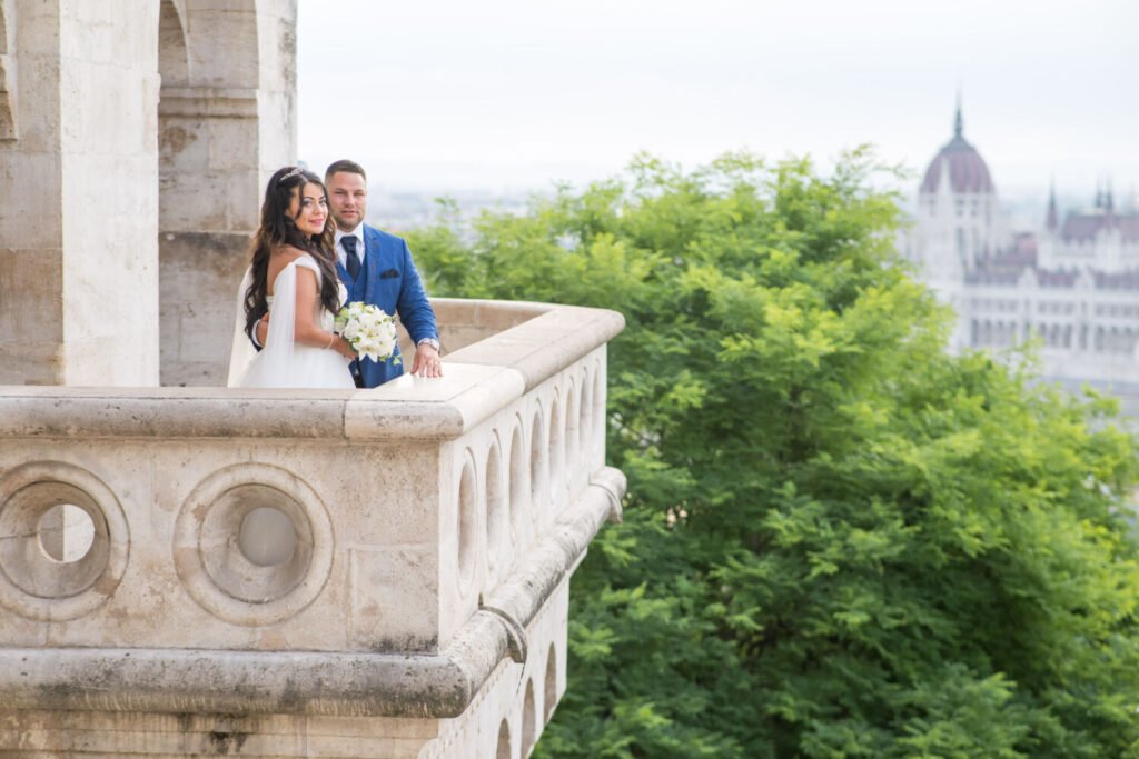 Esküvői művészi fotó a Halászbástya kilátópontjáról: fehér vállról lecsúszós menyasszonyi ruhás menyasszony és királykék öltönyös vőlegény a díszes kőkorlátnál állnak, kezükben fehér menyasszonyi csokor. A háttérben a Parlament kupolája és zöldellő fák látszanak, míg az előtérben a bástya jellegzetes kör motívumos kőfaragásai teszik még különlegesebbé ezt a panorámás esküvői felvételt.