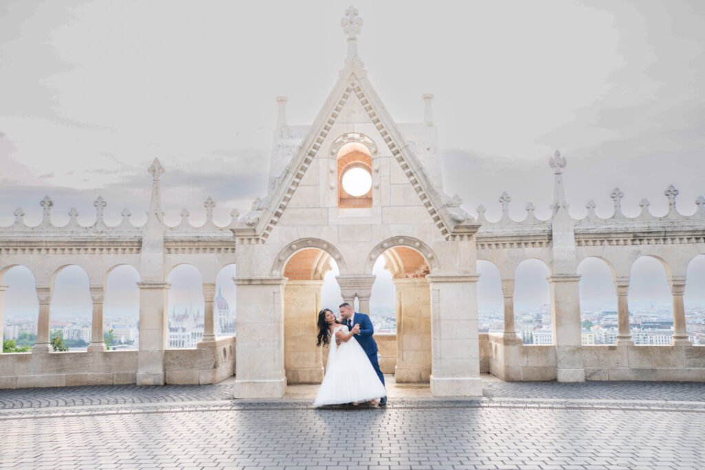 Esküvői fotóművész által megörökített táncmozdulat: kék öltönyös vőlegény keringőt táncol fehér tüllruhás menyasszonyával a Halászbástya gótikus kapuboltja alatt. A neogótikus építészet részletei, a fehér kőcsipkék és a háttérben feltűnő budapesti látkép harmonikus egységet alkotnak ebben a romantikus esküvői kompozícióban.