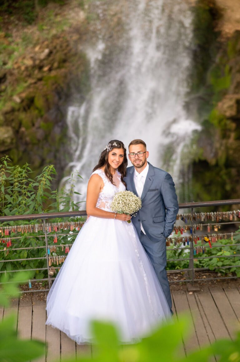 Esküvői kreatív fotózás a lillafüredi vízesés előtt – a menyasszony és a vőlegény boldogan pózol a híres lakatfalnál, háttérben a zuhatag látványával. A menyasszony elegáns fehér ruhában, kezében csodaszép virágcsokorral áll a mosolygó vőlegény mellett, aki klasszikus kék öltönyt visel. A természet varázsa és az érzelmek tökéletes harmóniája tükröződik ezen a romantikus esküvői portrén, amelyet egy profi esküvőfotós örökített meg.
