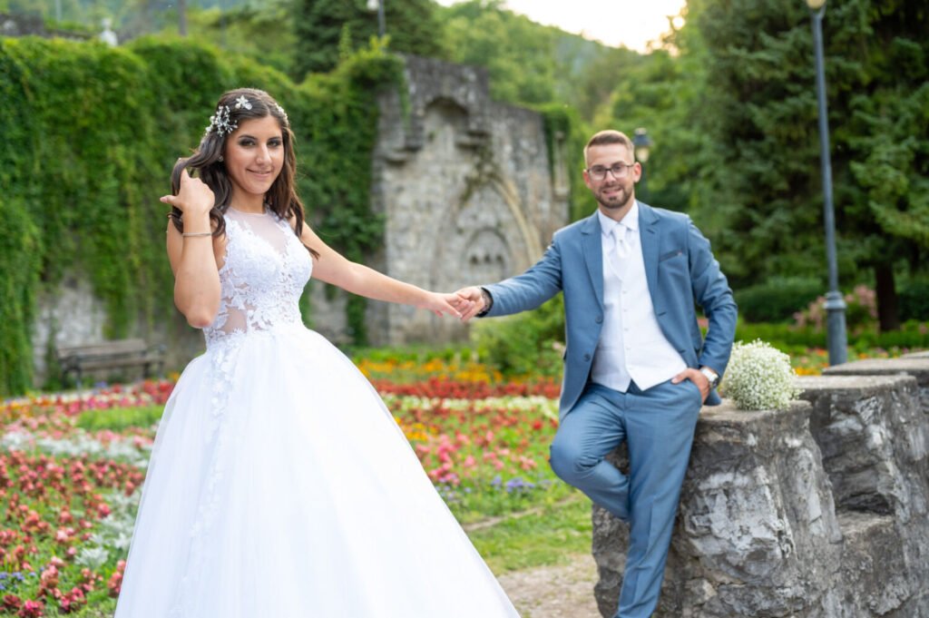 Esküvői kreatív fotóművész által megörökített játékos pillanat: csipke díszítésű fehér báli ruhás, virágos fejdíszes menyasszony és világoskék öltönyös vőlegény egy varázslatos kastélykertben, színes virágágyások és romantikus kőfal előtt. A pár kézenfogva, dinamikus pózban áll, miközben a háttérben borostyános falak és gótikus díszítőelemek teszik még különlegesebbé az esküvői portréfotózást.