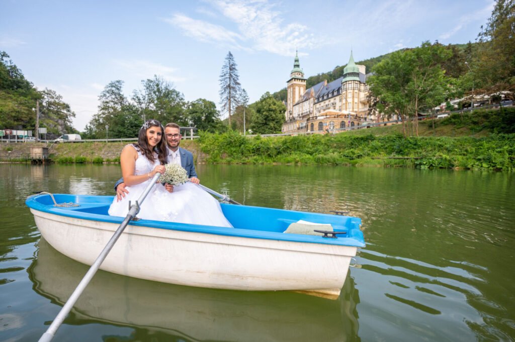 Esküvői riportfotós által megörökített panorámás kép: fehér-kék csónakban ülő ifjú pár a Hámori-tavon, háttérben a patinás lillafüredi Palotaszálló impozáns tornyaival. A fehér tüllruhás, virágdíszes menyasszony és világoskék öltönyt viselő vőlegény közös evezése során készült természetes pillanat, miközben a nyári égbolt és a zöldellő környezet keretezi ezt a különleges esküvői fotót.