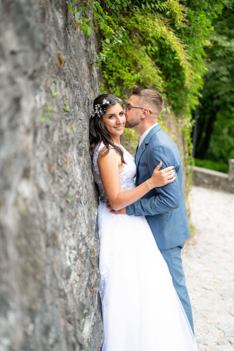 Esküvői fényképezés egy varázslatos szabadtéri helyszínen: a menyasszony egy ősi kőfalnak támaszkodva mosolyog a kamerába, míg a vőlegény gyengéden csókot lehel az arcára. A menyasszony elegáns fehér csipkeruhát visel, haját romantikus virágdísz ékesíti. A vőlegény világoskék öltönyben, fehér inggel és elegáns kiegészítőkkel öleli át szerelmét. A háttérben zöld növényzet és egy macskaköves ösvény teszi még idillibbé az esküvői fotózást. A profi esküvői fényképész tökéletesen megragadta a természetközeli, romantikus pillanatot.