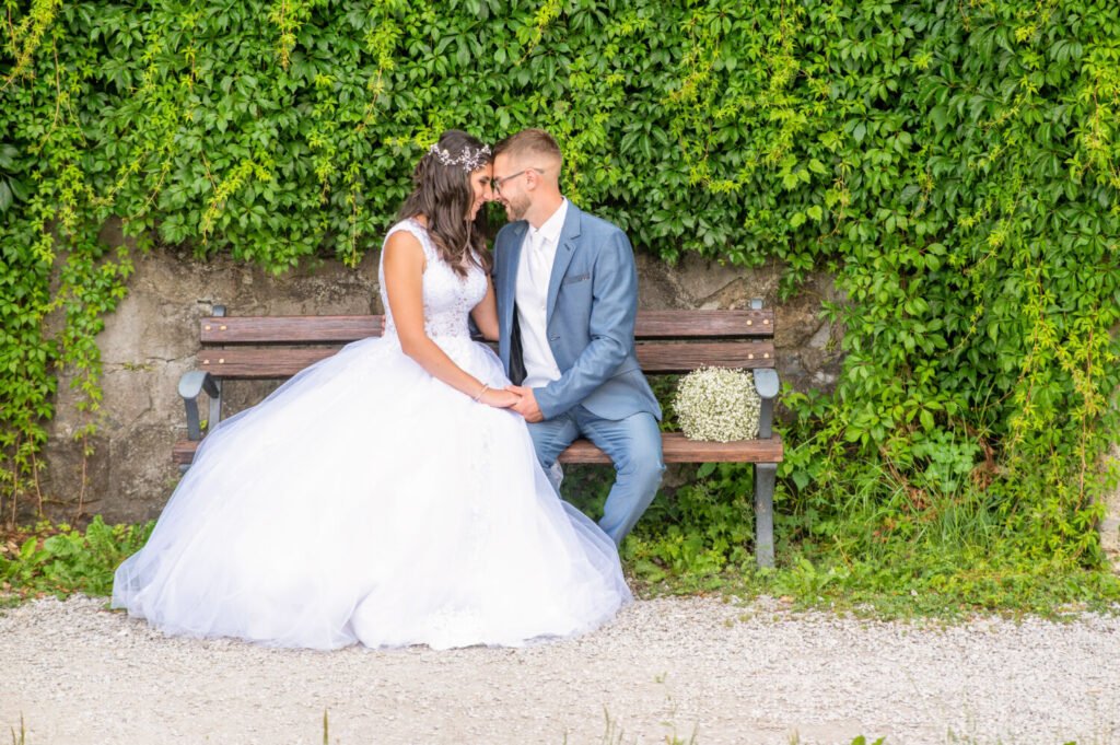 Esküvői fotóművész által megörökített intim pillanat: fehér tüll hercegnő ruhás menyasszony és világoskék öltönyös vőlegény egy hangulatos kerti padon ülve, zöld futónövényekkel borított fal előtt. A pár összeér, miközben a menyasszonyi ruha légiesen omlik alá, tökéletes művészi kompozíciót alkotva az esküvői portréfotózáson.