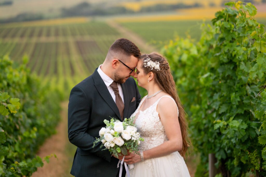 Esküvői fotóművész által megörökített romantikus pillanat a szőlősorok között: csipke díszítésű fehér menyasszonyi ruhás, ezüst hajdíszes menyasszony és fekete öltönyt, barna nyakkendőt viselő vőlegény gyengéden összeér homlokuk. A menyasszony kezében fehér rózsacsokor, a háttérben festői szőlőültetvények és dombok aranysárga színekben, tökéletes helyszínt biztosítva ennek a különleges esküvői portrénak.