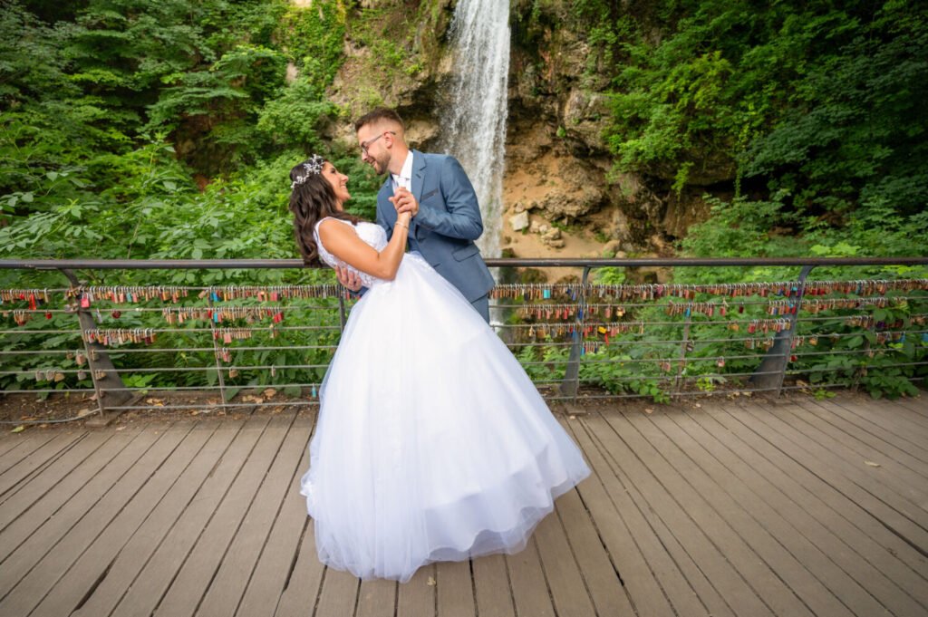 Esküvői történetmesélő fotográfus által megörökített táncmozdulat: a világoskék öltönyt viselő vőlegény kecsesen hátradönti fehér tüllruhás, virágkoszorús menyasszonyát egy fa hídon, színes szerelemlakatok sora és festői vízesés előtt. A dinamikus pillanat tökéletesen tükrözi az ifjú pár boldogságát, miközben a természet és a romantika jelképei veszik körül őket.