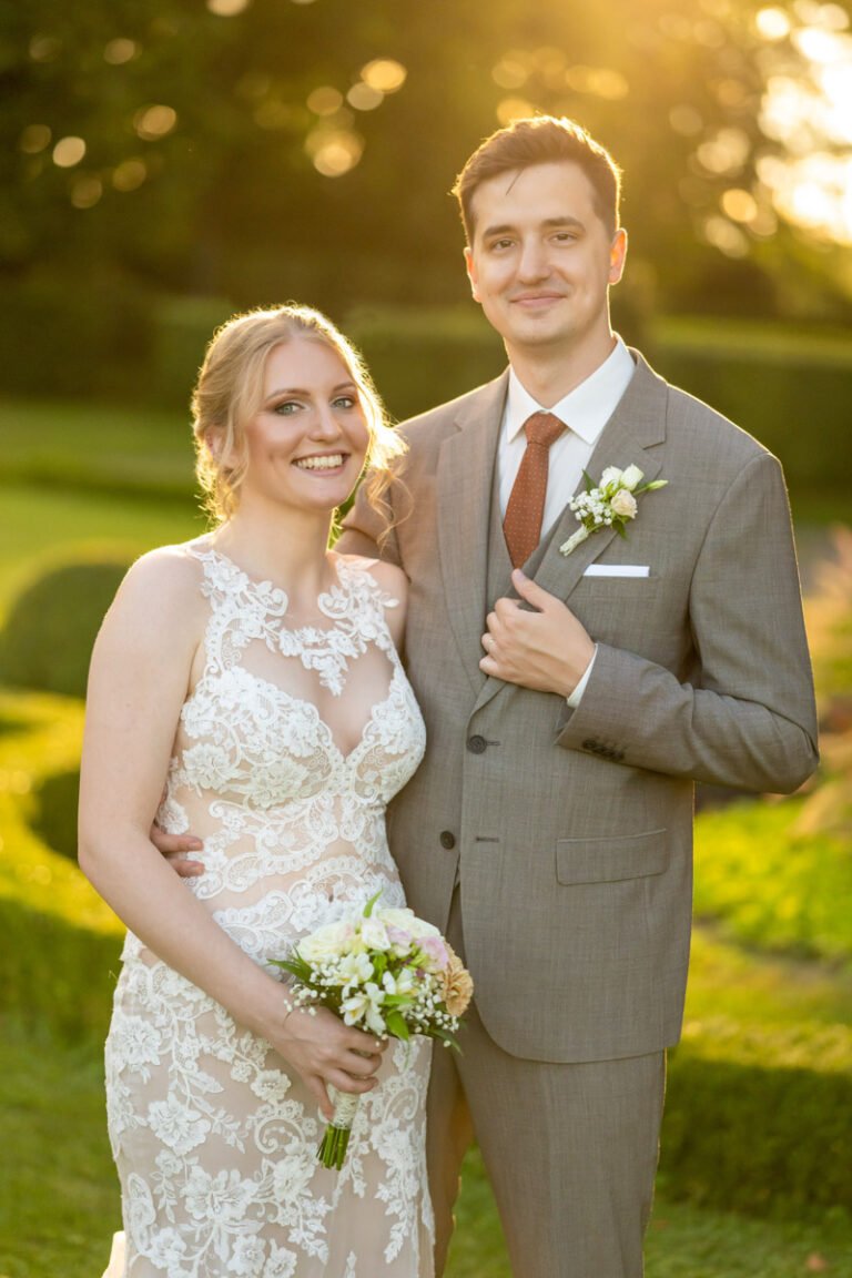 Esküvői fotós - Egy boldog ifjú pár áll a füzérradványi Károlyi-kastély gyönyörű kertjében, a lemenő nap aranyló fényében. A menyasszony csipkés esküvői ruhát visel, kezében romantikus virágcsokrot tart, miközben mosolyogva néz a kamerába. A vőlegény elegáns szürke öltönyt és barna nyakkendőt visel, mellkasán friss virágdísz tűnik fel.