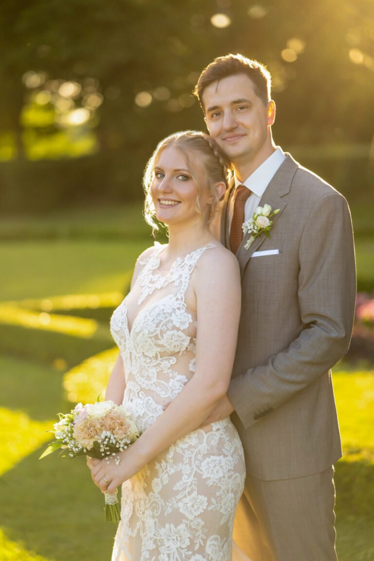 Egy szerelmes ifjú pár áll a füzérradványi Károlyi-kastély kertjében, a lemenő nap meleg fényében. A menyasszony finom csipkés esküvői ruhában mosolyog, kezében romantikus virágcsokorral, miközben vőlegénye gyengéden átöleli. A vőlegény elegáns szürke öltönyt és világos inget visel, mellkasán friss virágdísz látható. Az aranyóra fénye lágy és meghitt hangulatot kölcsönöz a fotónak, kiemelve az érzelmeket és az esküvői fotózás varázsát. Egy profi esküvői fényképész által megörökített természetközeli, elegáns és időtálló esküvői portré.