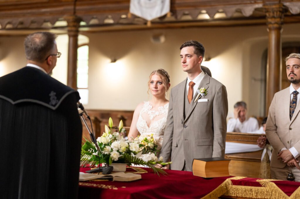 A menyasszony és a vőlegény meghitt pillanatban állnak az oltár előtt egy templomi esküvői szertartáson. A háttérben a lelkész vezeti a ceremóniát, miközben a pár komolyan és izgatottan figyel. Az elegáns virágdíszítés és a történelmi templombelső különleges hangulatot ad a pillanatnak. Az esküvői fényképész megörökítette a boldog és érzelmekkel teli momentumot, amely örökké emlékezetes marad.