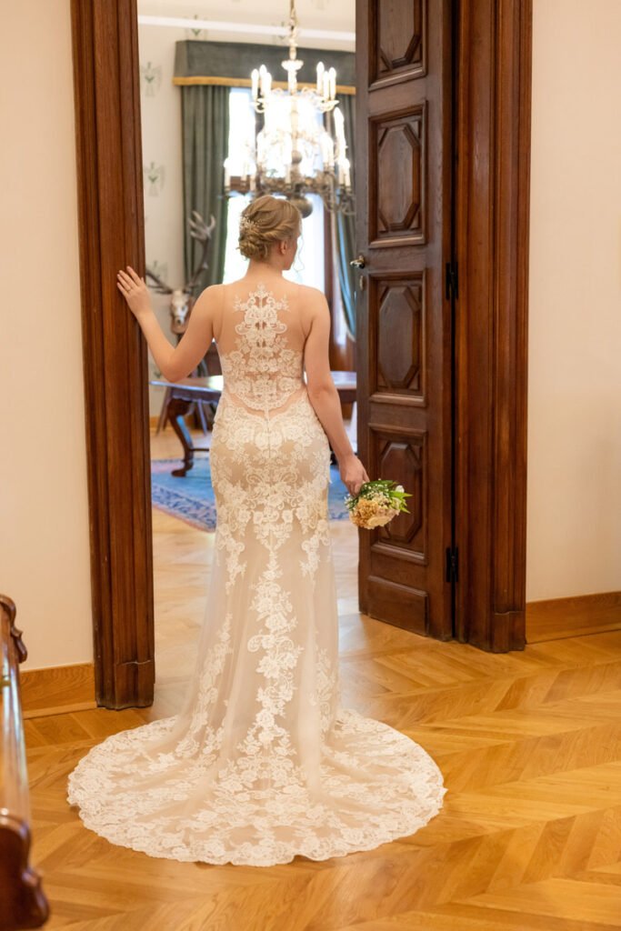 Hátulról fotózott menyasszony csipkés, uszályos esküvői ruhában, kezében egy finom virágcsokorral, ahogy egy impozáns faajtóban áll egy fényűző, díszes szalonban. A jelenetet egy profi esküvői fotós, nászfotós, lakodalmi fényképész, illetve menyegzői fotográfus örökítette meg a romantikus légkör és a meghitt pillanatok kihangsúlyozásáért.