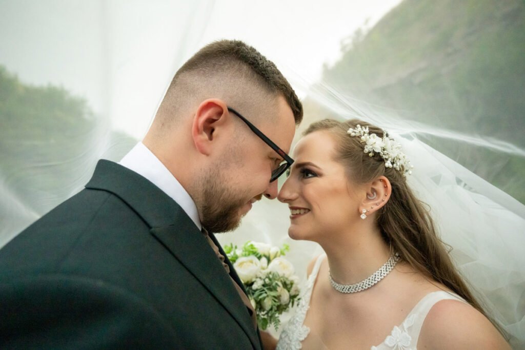 A menyasszony és a vőlegény egymás szemébe nézve mosolyognak, miközben a menyasszony fátyla finoman körülöleli őket. A vőlegény elegáns fekete öltönyt és barna nyakkendőt visel, míg a menyasszony csipkés esküvői ruhában, gyöngyös hajdísszel és gyönyörű ékszerekkel ragyog. A romantikus közeli portrét egy profi esküvői fotós, nászfotós, menyegzői fényképész és lakodalmi fotográfus örökítette meg.