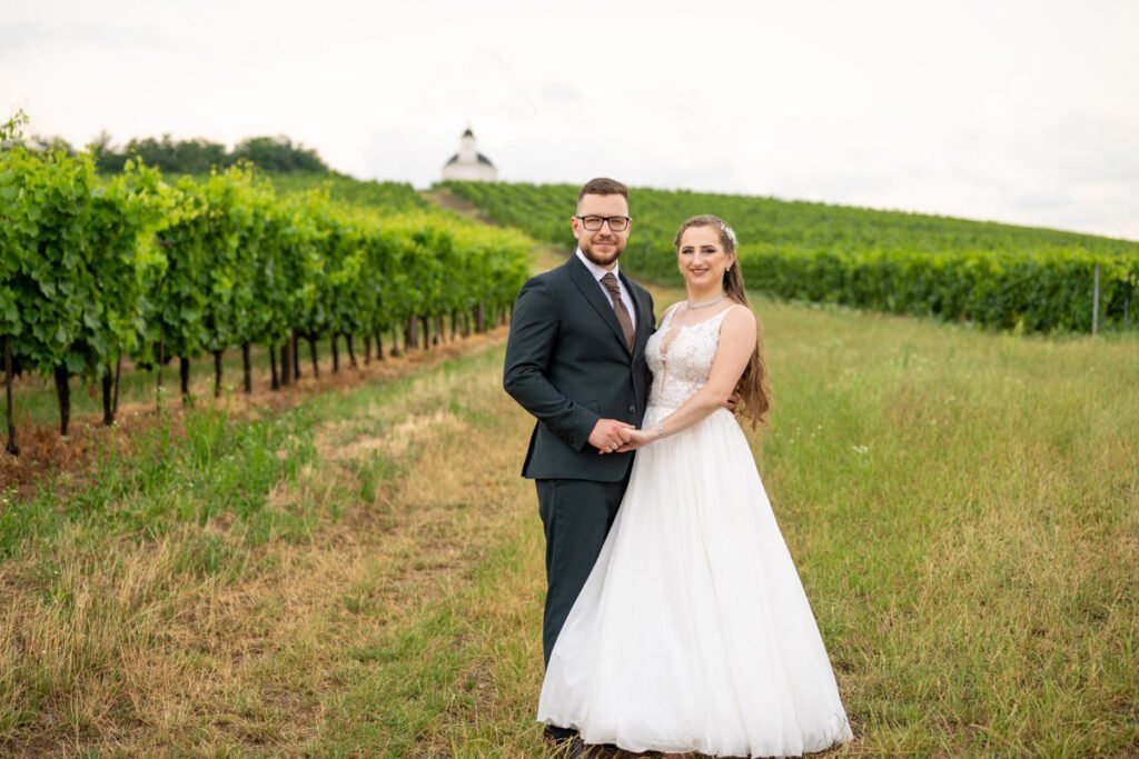 Esküvői fotográfus által készített romantikus kép egy boldog párról a tarcali szőlőültetvények között. A vőlegény elegáns fekete öltönyben, nyakkendővel és szemüvegben áll menyasszonya mellett, aki mesés csipkézett fehér esküvői ruhát visel. A pár kézen fogva, boldogan néz a kamerába, miközben a szőlőtőkék és a távoli dombtetőn álló Disznókő-kilátó festői hátteret biztosítanak. Esküvői fotózás vidéki hangulatban, természetközeli élménnyel és időtlen szépséggel.