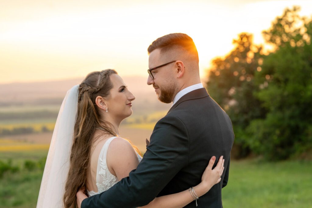 Esküvői fotózás varázslatos pillanata, ahol a menyasszony és a vőlegény szerelmesen néznek egymás szemébe a naplemente aranyló fényében. A menyasszony elegáns csipkeruhát és fátylat visel, míg a vőlegény stílusos öltönyben öleli párját. A háttérben festői táj és aranyló égbolt teszi tökéletessé a romantikus hangulatot. Profi esküvői fényképész által megörökített meghitt pillanat, amely örök emléket nyújt az ifjú pár számára. Kreatív esküvőfotózás szabadtéri helyszínen, természetes fénnyel és érzelmekkel teli kompozícióval.