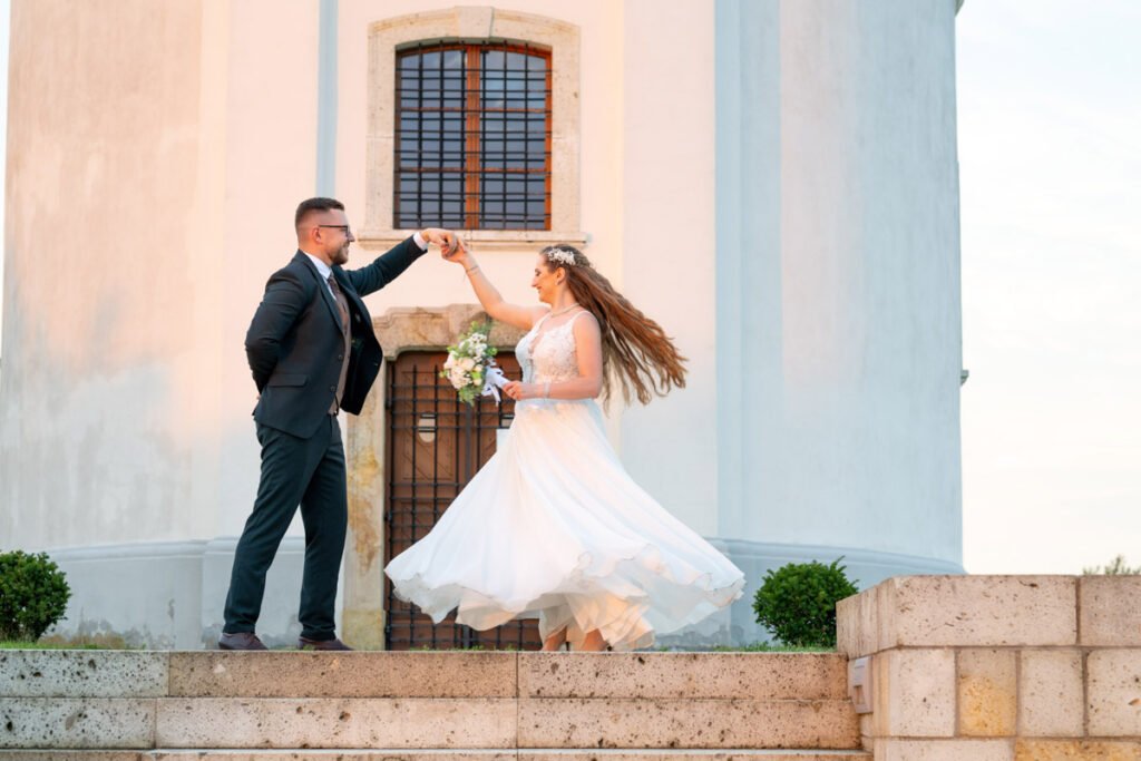 Romantikus esküvői fotó, ahol a vőlegény és a menyasszony önfeledten táncol a templom lépcsőjén egy varázslatos naplementében. A menyasszony hófehér, légies ruhája kecsesen lebeg a mozdulatokkal, kezében egy csodás virágcsokorral. A vőlegény elegáns fekete öltönyt visel, és mosolyogva vezeti párját a táncban. A háttérben a történelmi épület impozáns homlokzata emeli ki az esküvői fotó művészi hangulatát. Kreatív esküvői fotózás, amely a szerelem és az öröm pillanatait örökíti meg profi esküvőfotós által.