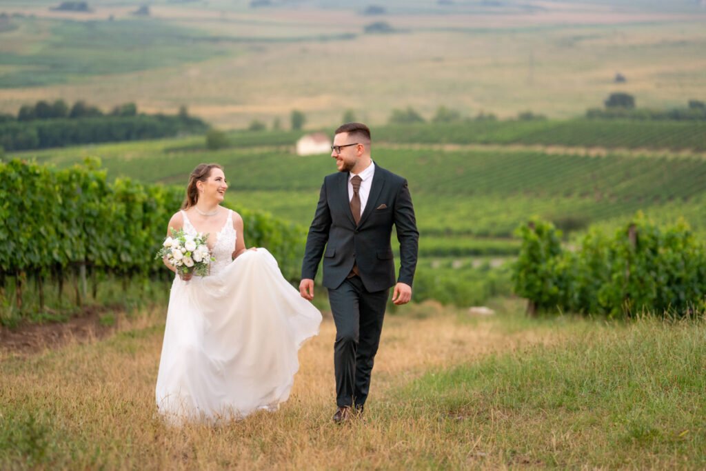 Esküvői kreatív fotózás a természetben: a boldog ifjú pár kézen fogva sétál egy gyönyörű, zöld szőlőhegy lankáin. A menyasszony hófehér, csipkézett esküvői ruháját finoman megemeli, miközben mosolyogva tekint szerelmére. Kezében elegáns fehér rózsákból és zöld növényekből álló menyasszonyi csokor díszeleg. A vőlegény stílusos fekete öltönyt visel, barna nyakkendővel és zsebkendővel kiegészítve. A háttérben festői táj és végtelen szőlősorok láthatók. Esküvői fotográfus által készített természetes és érzelemdús fénykép az igaz szerelem pillanatáról.