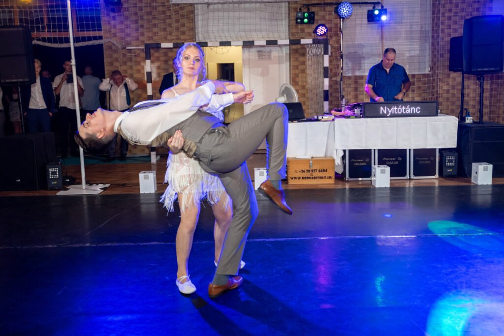Esküvői fényképész által megörökített dinamikus pillanat a lakodalom nyitótáncáról. Egy elegáns ruhába öltözött pár mutat be egy lenyűgöző tánckoreográfiát, ahol a nő magabiztosan tartja partnerét egy merész emelés során. A háttérben DJ pult és táncoló vendégek láthatók, akik az ünnepi pillanat részesei. A fények és a tükörsima táncparkett egyedi hangulatot kölcsönöz az esküvői fotónak.