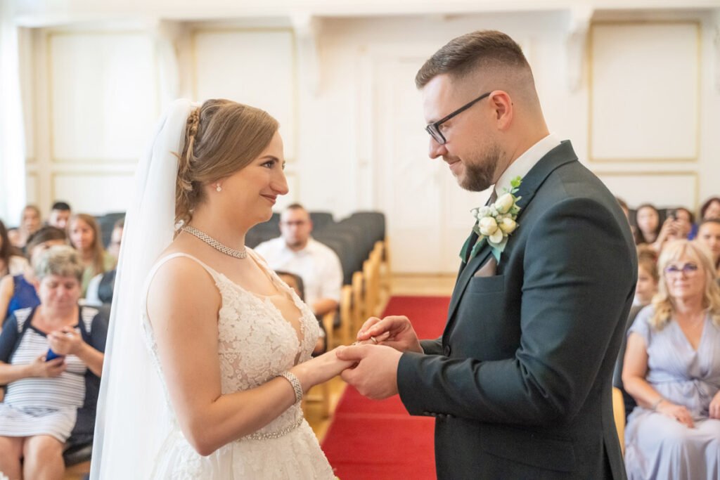 Esküvői fotográfus által megörökített gyűrűhúzás a polgári szertartáson: a menyasszony és a vőlegény szerelmes pillantással néznek egymásra, miközben a vőlegény óvatosan az ujjára húzza a jegygyűrűt. A menyasszony gyönyörű fehér csipkeruhát és csillogó nyakéket visel, fátyla lágyan omlik a vállára. A vőlegény elegáns sötét öltönyben, fehér ingben és zöld-fehér virágkitűzővel áll előtte. A háttérben a vendégek meghatódva figyelik az esküvői ceremónia varázslatos pillanatait. Profi esküvői fényképész által készített kép, amely tökéletesen visszaadja a nagy nap érzelmeit és a házasságkötés örömét.