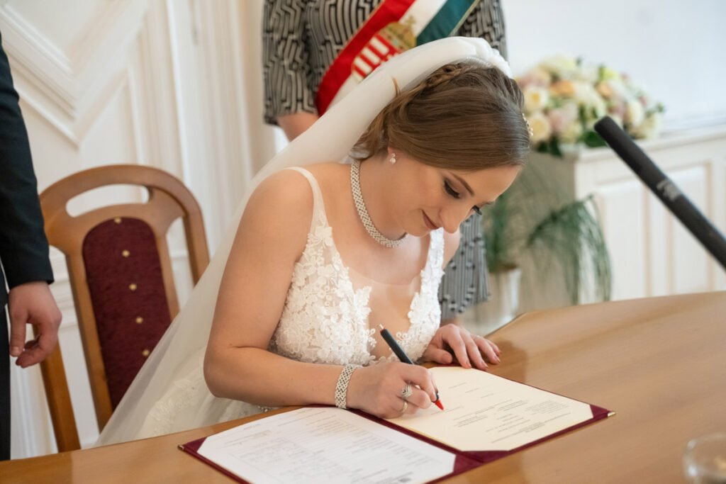 Esküvői fényképész által megörökített különleges pillanat: a menyasszony boldog mosollyal írja alá a házassági anyakönyvi kivonatot az esküvői ceremónián. Fehér csipkés menyasszonyi ruhában, elegáns fátyollal és csillogó ékszerekkel ragyog az asztalnál, miközben az anyakönyvvezető nemzeti szalaggal a háttérben áll. Az esküvőfotó a meghitt és ünnepélyes pillanatok szépségét örökíti meg, tökéletes példa a professzionális esküvői fényképezés jelentőségére. Esküvői fotográfus által készített érzelemdús és jelentőségteljes kép.