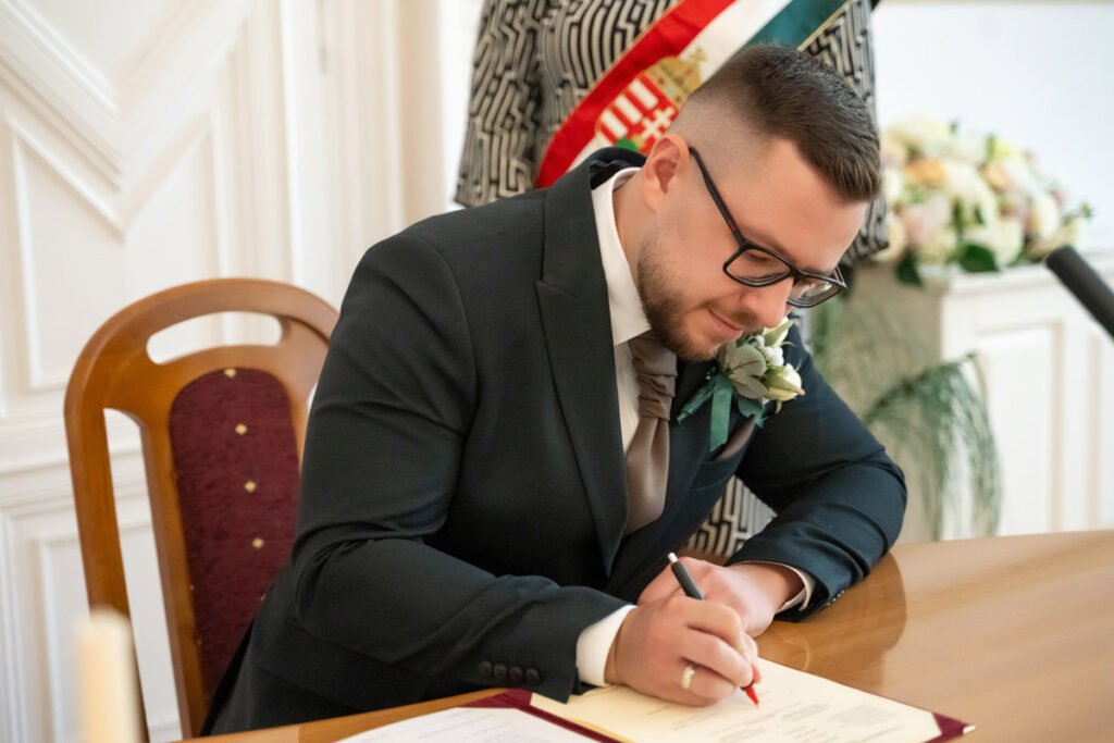 Esküvői fotográfus által megörökített meghitt pillanat: a vőlegény elegáns fekete öltönyben és fehér virágos kitűzővel írja alá a házassági anyakönyvi kivonatot. Az esküvői fotózás egyik legfontosabb dokumentációs momentuma, amely a ceremónia hivatalossá tételét örökíti meg. A háttérben az anyakönyvvezető nemzeti szalaggal, valamint fehér és pasztell virágdekoráció látható. A profi esküvői fényképezés segítségével a pillanat időtálló emlékké válik, amely a menyegző egyik kulcsfontosságú mozzanatát rögzíti.