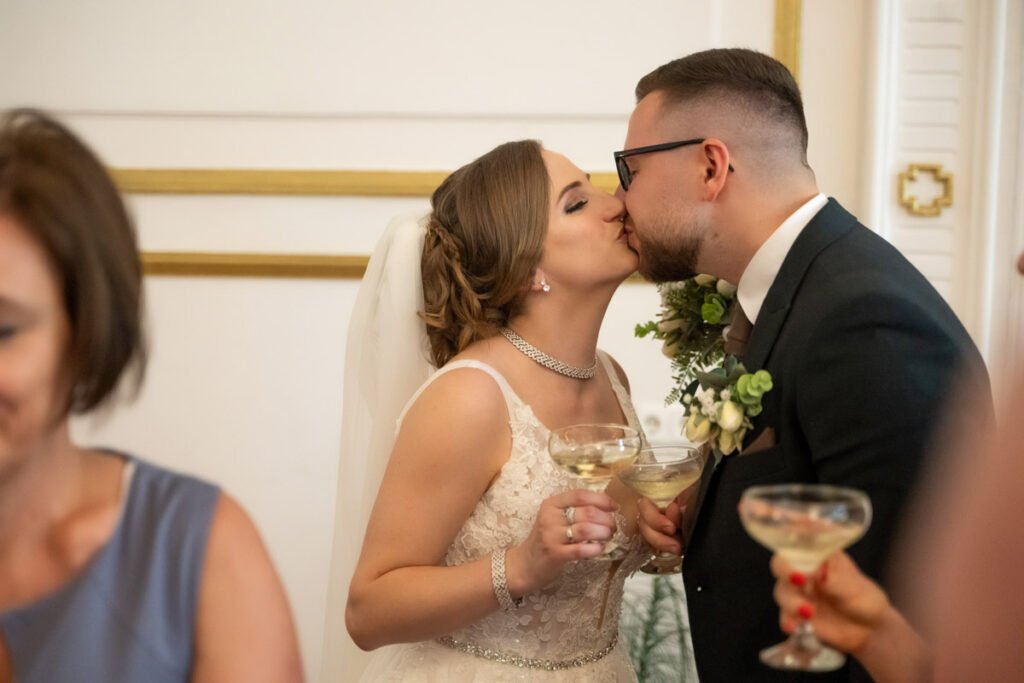 Esküvői fényképezés során megörökített romantikus pillanat: az ifjú pár boldog csókot vált az anyakönyvi ceremónia után, miközben pezsgőspoharat tartanak a kezükben. A menyasszony elegáns fehér csipkeruhában, csillogó ékszerekkel és fátyollal tündököl, míg a vőlegény sötét öltönyben, fehér virágos kitűzővel áll mellette. A háttérben vendégek és ünnepi dekorációk láthatók. A profi esküvői fotós tökéletesen megragadta az érzelmekkel teli pillanatot, amely az esküvői fotózás egyik legmeghatóbb jelenete.